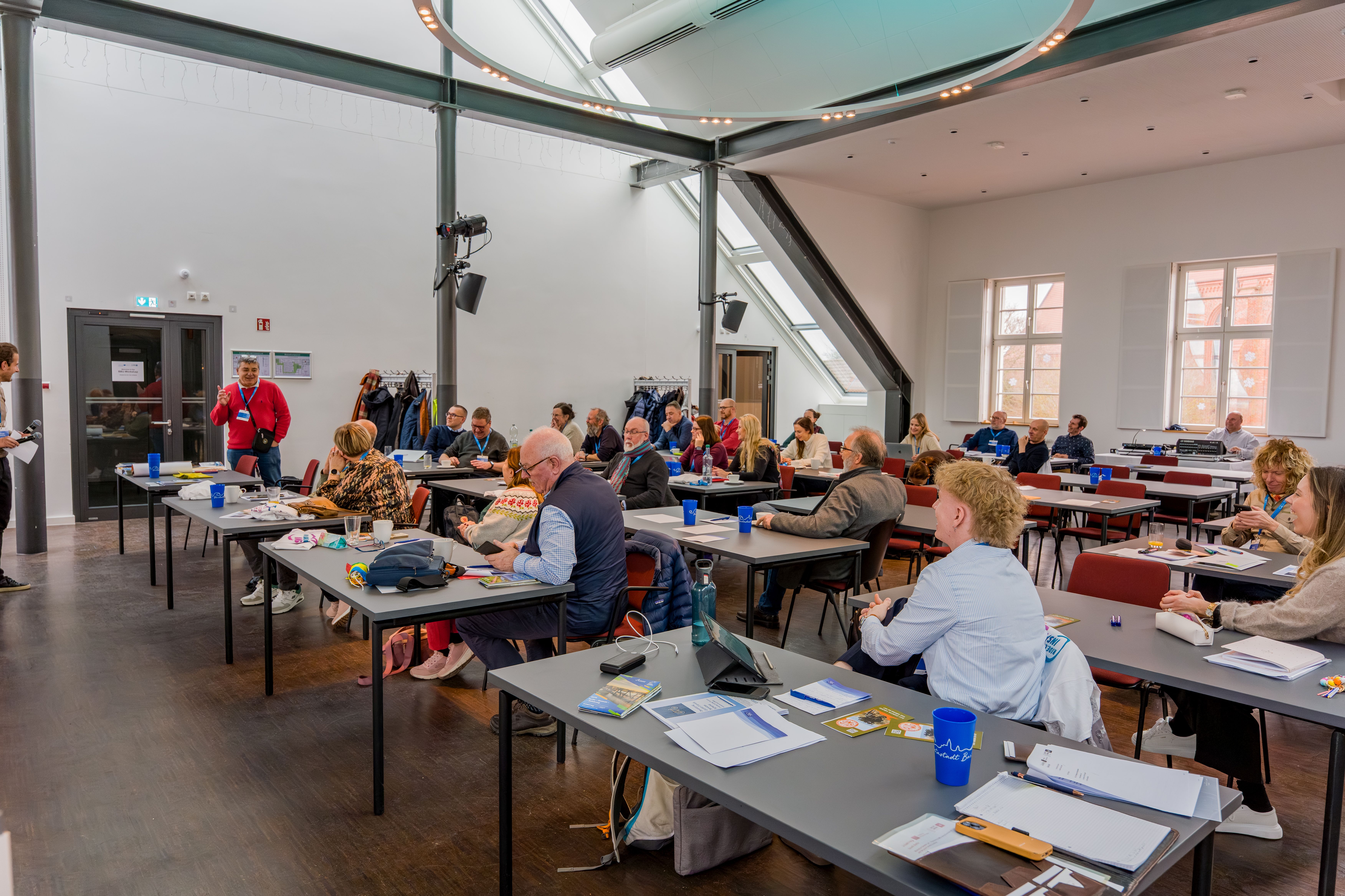 Menschen sitzen in einem Saal zusammen an einzelnen Tischen und hören einen Vortrag